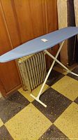 Full view of ironing board standing upright showing metal legs and blue cover