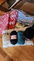 Photo showing 4 skeins of yarn in cream, black, and teal colors on top of a packaged item, with red and blue patterned linens in the background.