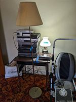 Full view of brass tone floor lamp next to stereo system and chair, showing lamp standing on the floor with beige fabric top shade and hobnail milk glass shade below.