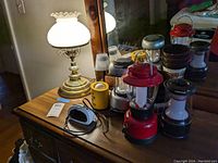 Brass table lamp with white milk glass hobnail shade lit on a wooden dresser with Coleman style camping lanterns and a travel alarm clock.