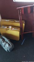 Photo showing two yellow Sears suitcases stacked upright next to wooden luggage rack with reddish-brown finish and tan leather straps.
