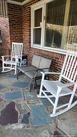 Overview of two white painted wood rocking chairs and one metal mesh porch glider on stone porch beneath window.