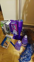 Swiffer WetJet mop with accessories including cleaning solution bottles, blue mop head, and dustpan with brush on a wood floor