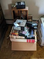 Boxes and bag of assorted VHS tapes, cassettes, CDs, and a black VHS rewinder visible on top of the pile.