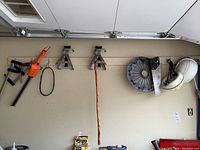 Wide view of garage wall showing car stands, hydraulic jack, clamps, and hedge trimmer hanging on hooks.