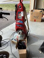 Full front view of the red upright Bissell ProHeat carpet cleaner showing power cord, hose attachment, and small box of accessories nearby.