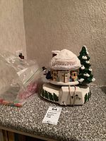 Ceramic Christmas dome music box featuring a snowy roof with 'Happy Holidays' message, snowman figure, and a green Christmas tree next to it, with two bags of Christmas lights beside it on countertop.