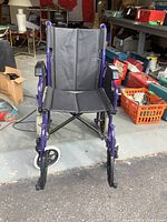 Frontal view of the purple folding wheelchair showing black fabric seat and backrest with armrests and small front wheels.