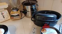 Photo shows Oster blender with clear glass jar on beige base next to a white mechanical kitchen scale. Nearby are a black roasting pan, a loose base round flan sandwich pan with label, and stacked stainless steel pots with lids.