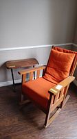 Wood frame rocking chair with orange cushions and wooden side table beside it in a corner.