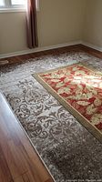 Rug with red center leaf motif and ornate beige and brown outer pattern on wood floor near window with curtain.
