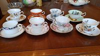 Photo shows all eight tea cups with matching saucers displayed on a wooden table. Various floral and seasonal patterns visible.