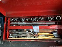 Top view of the open toolbox showing an organized socket set, multiple hand tools including vice grips, pliers, and blades inside a red metal toolbox.