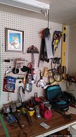 Tools mounted on pegboard wall and assorted tools on wooden work surface beneath showing a variety of hand tools, measuring tape, clamps, saw blades, and power tool case.