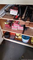 Overview of cupboard interior showing shelves with woodworking tools, hardware boxes, sanding pads, and paint can.