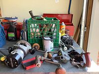 Various vintage hand tools on metal table including crank hand drill, and small red hand plane, green plastic crate with tools inside, spray can of tire foam and oil containers, red metal toolbox in background