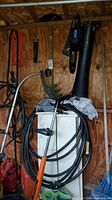 Wide view of items hanging on wall and placed on cabinet including trimmers, chainsaw, gas cans, hose.