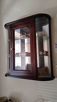 Front angled view of the wall display cabinet showing dark wood frame, glass front door with a brass knob, and curved glass sides.