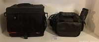 Front view showing black Canon Lowepro camera bag with red logo and black Optex camcorder bag with leather-like texture, both on shelf