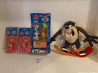 Front view of four Looney Tunes items including two safety flashers in packaging, Bugs Bunny candy handler in packaging with Skittles candy, and Taz plush on skateboard.
