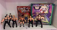 Front view of 6 loose wrestling figures in black NWO attire and 2 boxed items, including the Stone Cold Steve Austin figure and wrestling ring playset boxed and sealed.