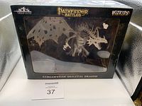 Front view of the sealed black box containing the Gargantuan Skeletal Dragon miniature figure, showcasing the unpainted skeleton figure through a clear window.