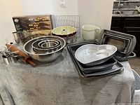 Wide shot showing full assortment of bakeware items on a towel-covered table including pans, bowls, rolling pins, measuring tools, glass pie plates, and cooling racks under neutral lighting.