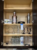 Shelving unit with assorted barware items including cocktail shakers, bottles, martini glasses, photo coaster box, mugs, stein, tumblers, and tools.