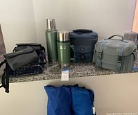 Full display of camping accessories including Stanley thermoses, Bushnell binocular cases, lunch box, blanket, and other items on countertop.