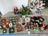 Wide shot of assorted Christmas decorations including boxed Lenox ornaments, figurines, small tree, stockings, and other holiday décor items arranged on a floor surface.