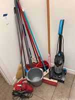 Full view of cleaning tools assortment including brooms, mops, bucket, Bissell PowerForce sweeper and Dirt Devil handheld sweeper.