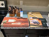 Photo of two wooden knife blocks with assorted knives including Wüsthof brand, wooden cutting board, meat tenderizer, boxed Grillworks grilling set, and boxed KüchenStolz cutlery set on a kitchen counter.