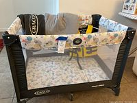 Graco Pack N Play playpen with animal print fabric and mesh sides, padded mattress, carry bag visible behind playpen. Airhead youth life vest in yellow and black partially inside the playpen.