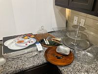 Assortment of party servingware on kitchen counter including glass platters, wooden platters, three-tier serving tray, glass dome, cheese slicer and other utensils.