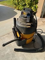 Full view of the yellow and black wet/dry vacuum with hoses attached, sitting on a concrete driveway with some dirt visible on the unit.