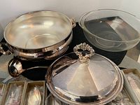 Silver plated serving tureen with ornate handles and lid, includes clear glass dish insert and ladle on black cloth.