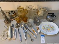 Wide view of assorted vintage kitchenware including pitchers, teapots, utensils with white handles, small electric cutter, two amber glass cups, stainless steel kettle, and serving platter.