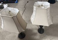Photo showing two black base table lamps with white fabric shades on a beige carpet.
