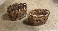 Two oval woven baskets, medium brown color, placed on carpeted floor, both with handles on short ends.
