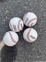 Four Rawlings Official League baseballs on asphalt with visible dirt and scuff marks, showing signs of use.