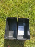 Top-down photo of the two open black marine battery boxes placed on grass, showing their interiors with one box containing papers.