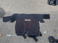 Front view of Bohn ButtSaver protective gear lying on pavement showing adjustable Velcro and strap fasteners, black with red stitching.