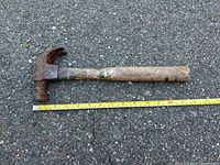 Photo showing a claw hammer on concrete with a measuring tape for scale, hammer head has rust and wooden handle is weathered.