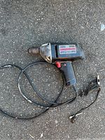 Black and silver Craftsman electric drill with cord and plug on asphalt.