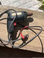 Black corded electric drill with rust-colored chuck and red trigger switch on wooden surface, power cord and red plug adapter visible.