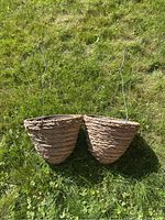 Two brown wicker hanging baskets laid on grass showing their woven texture and attached metal wire hooks for hanging.