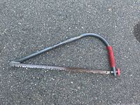 Photo of entire angled bow saw laying on asphalt surface, showing metal frame, 21-inch serrated blade, and red grip handle.