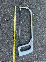Manual hacksaw with slotted metal frame and black blade; measuring tape beside it for scale.