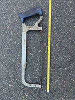 Full view of a metal hand saw with blue handle placed on asphalt surface next to a yellow measuring tape.