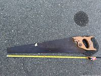 Photo of a traditional hand saw with wooden handle and a long metal blade on asphalt with a measuring tape for scale.